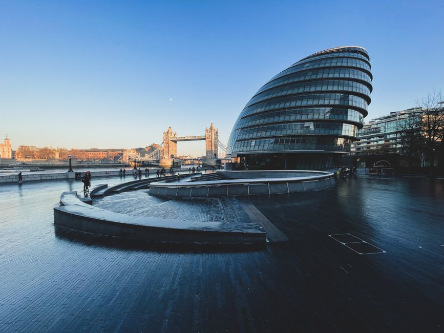 Wide-angle view of London Bridge station's outdoor area during daylight, showing a modern, curved glass building with multiple floors, reflecting the blue sky. In the foreground, a paved open space wi