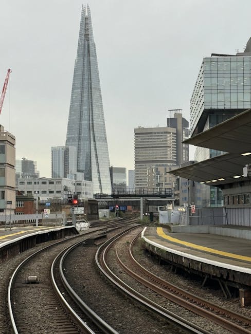 Wide-angle view of London Bridge station's outdoor area during daylight, showing a modern, curved glass building with multiple floors, reflecting the blue sky. In the foreground, a paved open space wi
